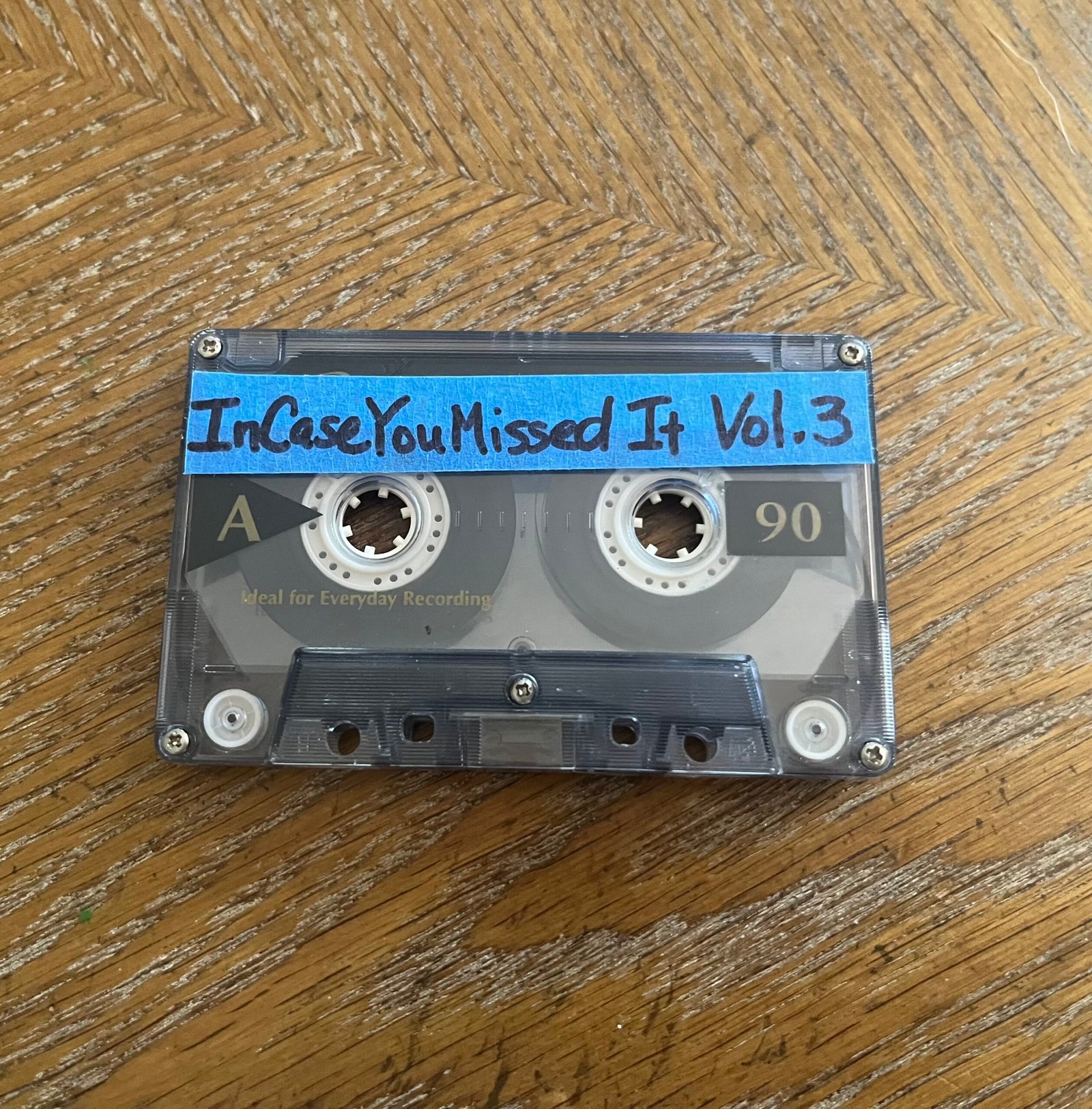 A cassette tape sits on a brown wooden table top. The tape is market with a blue handmade label with “In Case You Missed It Vol. 3’ written on it in black sharpie marker.